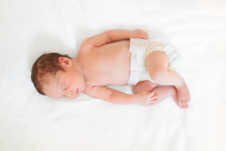 Baby in diaper in white sunny bedroom. Newborn child relaxing in bed. Nursery for young children. Textile and bedding for kids. Family morning at home. New born kid in nappy.の写真素材