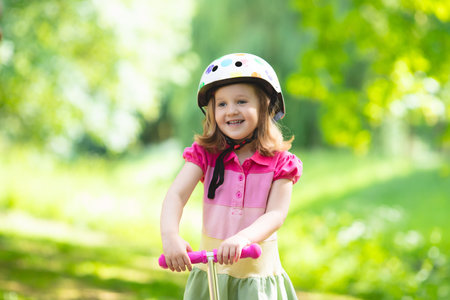 Little child learning to ride a scooter in a city park on sunny summer day. Cute preschooler girl in safety helmet riding a roller. Kids play outdoors. Active leisure and outdoor sport for children.の写真素材