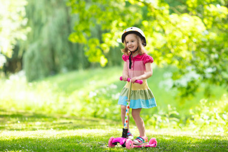 Little child learning to ride a scooter in a city park on sunny summer day. Cute preschooler girl in safety helmet riding a roller. Kids play outdoors. Active leisure and outdoor sport for children.の写真素材