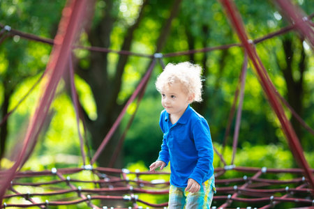 Active little child playing on climbing net and jumping on trampoline at school yard playground. Kids play and climb outdoors on sunny summer day. Funny boy on nest swing at preschool sport center.の写真素材
