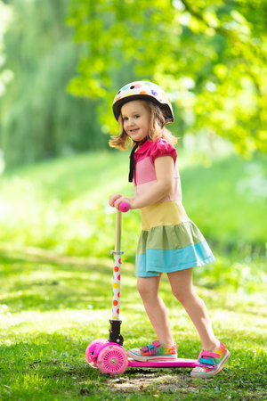 Little child learning to ride a scooter in a city park on sunny summer day. Cute preschooler girl in safety helmet riding a roller. Kids play outdoors. Active leisure and outdoor sport for children.の写真素材