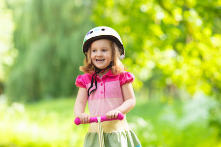 Little child learning to ride a scooter in a city park on sunny summer day. Cute preschooler girl in safety helmet riding a roller. Kids play outdoors. Active leisure and outdoor sport for children.の写真素材