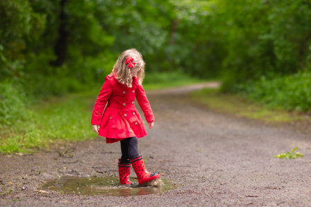 Little girl playing in rainy summer park. Child with red ladybug umbrella, waterproof coat and boots jumping in puddle and mud in the rain. Kid walking in autumn shower. Outdoor fun by any weather.の写真素材