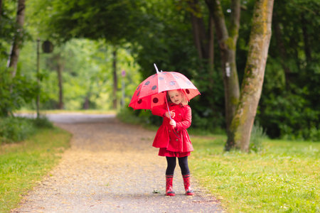 Little girl playing in rainy summer park. Child with red ladybug umbrella, waterproof coat and boots jumping in puddle and mud in the rain. Kid walking in autumn shower. Outdoor fun by any weather.の写真素材