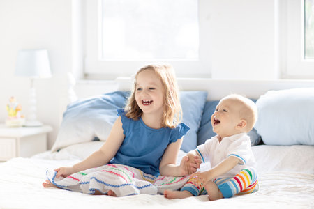Little girl and baby boy, brother and sister playing in parents bed. Family with children in the morning. Kids play in white bedroom. Nursery crib bedding and textile for young child. Siblings love.の写真素材