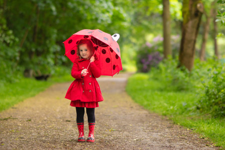 Little girl playing in rainy summer park. Child with red ladybug umbrella, waterproof coat and boots jumping in puddle and mud in the rain. Kid walking in autumn shower. Outdoor fun by any weather.の写真素材