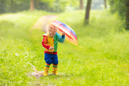 Little boy playing in rainy summer park. Child with colorful rainbow umbrella, waterproof coat and boots jumping in puddle and mud in the rain. Kid walking in autumn shower. Outdoor fun by any weatherの写真素材