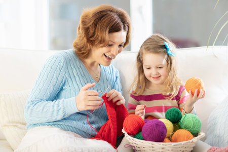 Mother and daughter knitting woolen scarf. Mom teaching child to knit. Crafts and hobbies for parents and children. Toddler girl kid with wool yarn in a basket. Knitted clothing for family with children.の写真素材