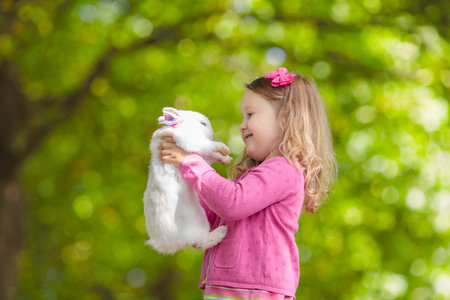 Children play with rabbit. Laughing child at Easter egg hunt with white pet bunny. Little toddler girl playing with animal in the garden. Summer outdoor fun for kids with pets.の写真素材