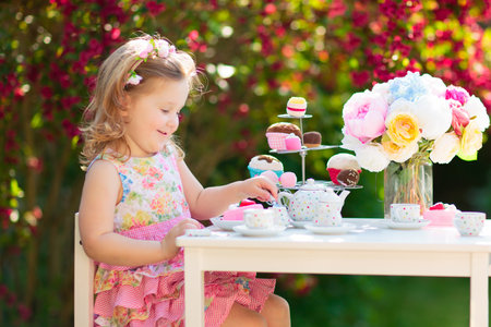 Tea garden party for kids. Child birthday celebration. Little boy and girl play outdoor drinking hot chocolate and eating cake. Children eat sweets. Kid event with toy dish and flower decoration.の写真素材