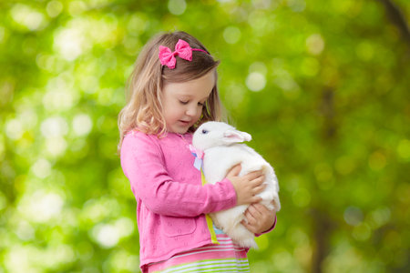 Children play with real rabbit. Laughing child at Easter egg hunt with white pet bunny. Little toddler girl playing with animal in the garden. Summer outdoor fun for kids with pets.の写真素材