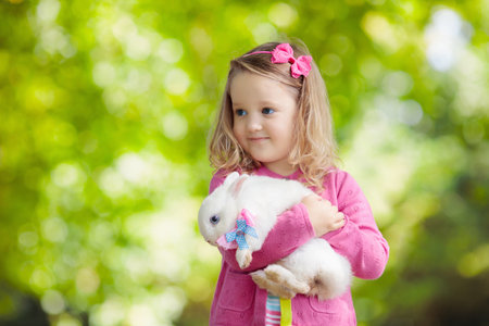 Children play with real rabbit. Laughing child at Easter egg hunt with white pet bunny. Little toddler girl playing with animal in the garden. Summer outdoor fun for kids with pets.の写真素材