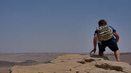 Young man with backpack sitting on cliffs edge and looking at the desertの写真素材