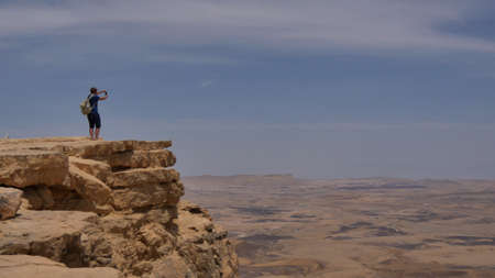 Young man standing on cliff edge and taking panoramic photo of the desert on his phoneの写真素材