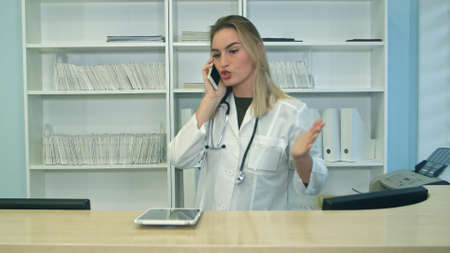 Young stressed woman yelling and gesturing while speaking on telephone at hospital receptionの写真素材