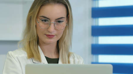 Concentrated female doctor in glasses working on laptop at reception deskの写真素材