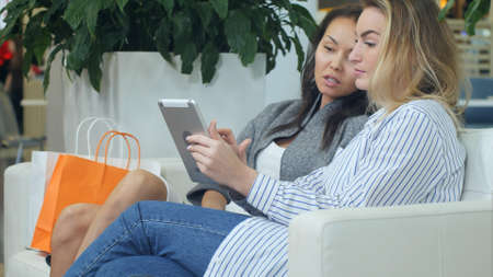 Two female friends having fun while shopping in a mall, having a break, using digital tablet and talkingの写真素材