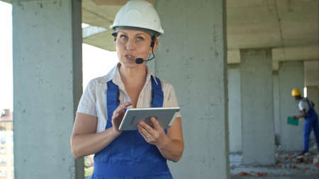 Female construction engineer reading plans using digital tablet and talk to workers through internal...