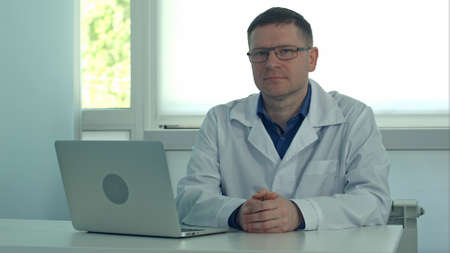 Mature male doctor sitting at desk with laptop and looking at camera in his clinic officeの写真素材