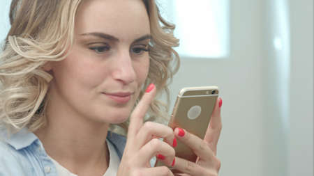 Happy young blond woman is sitting in a beauty salon and using a smartphoneの写真素材