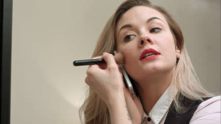 Young beautiful woman applying makeup on face with brush, having phone call, being late
