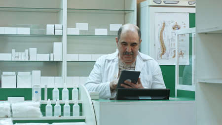 Senior pharmacist with mustache working on tablet pc checking medicine in pharmacyの写真素材