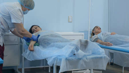 Two female patients on drips lying on beds in hospital ward being checked by two doctorsの写真素材