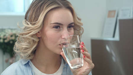 Gorgeous young woman is keeping healthy by drinking a glass of water indoors and smilingの写真素材
