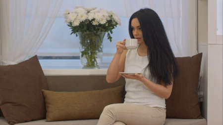 Young brunette sitting on sofa drinking tea from cup in a officeの写真素材