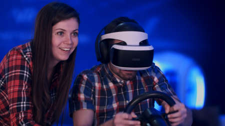 Pretty smiling girl watching her boyfriend playing racing video game in virtual reality headset