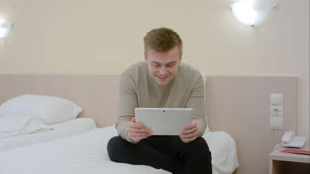 Young man just coming to a hotel room, sitting on bed and having video chat on tabletの写真素材