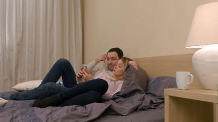 Loving couple sitting on the bed, using smartphone, discussing new photosの写真素材
