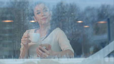 Beautiful girl using phone while drinking coffee in a cafe viewed through windowの写真素材
