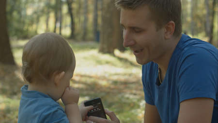 Father helps crying son to use cell phone in park.の写真素材