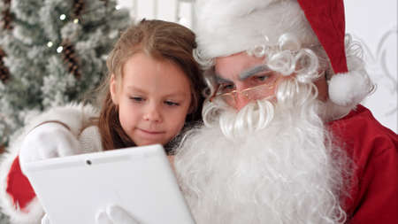 Santa Claus taking Christmas selfie on tablet with cute little girl sitting on his lapの写真素材