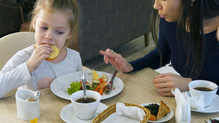 Beautiful brunette mother persuading her young blond daughter to eat vegetable salad in cafe.の写真素材