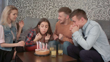 Attractive teen girl celebrates her birthday with friends at home and blows out the candles on cake and drinks orange juceの写真素材