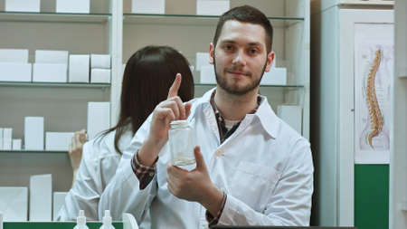 Young malepharmacist holding a white blank bottle of pills, promoting medicine, while his colleague workingの写真素材
