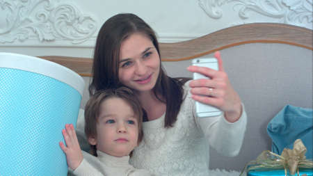 Young mother and happy son holding Christmas presents taking selfies with phoneの写真素材