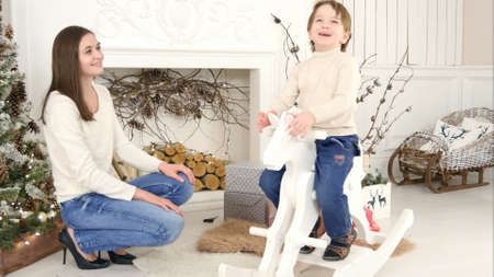 Little boy riding wooden rocking horse and talking to his mother sitting near the Christmas treeの写真素材