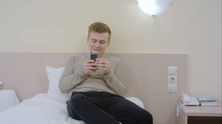 Handsome young man using smartphone and laughing in the bedroom at homeの写真素材