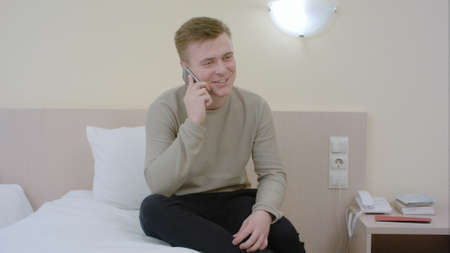 Man talking and laughing on his smartphone seated on a bedの写真素材