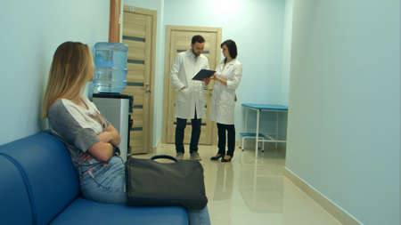 Worried woman patient waiting in hospital hall while two doctors discussing diagnosisの写真素材