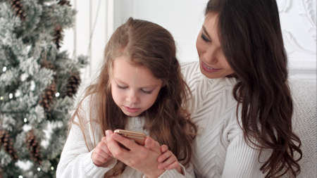 Young mother teaching her daughter how to use smartphone in Christmas decorated roomの写真素材