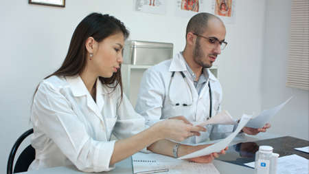 Two busy doctors working with papers and xray images, answering phone calls, prescribing pills at their deskの写真素材