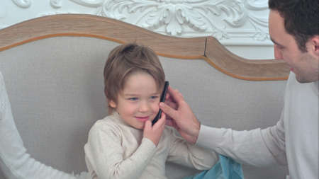 Little boy sitting on the sofa talking on the phone passed to him by his fatherの写真素材