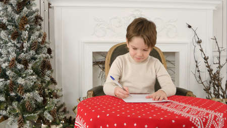 Serious little boy writing letter to Santa sitting at the table near the Christmas treeの写真素材