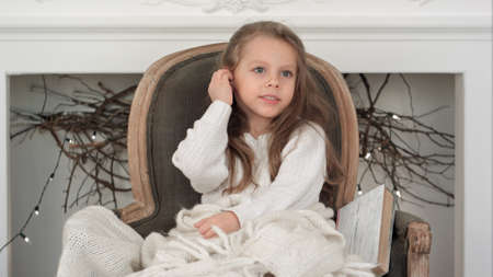 Little smiling girl in white sweater sitting on a chair wrapped in white Christmas blanketの写真素材