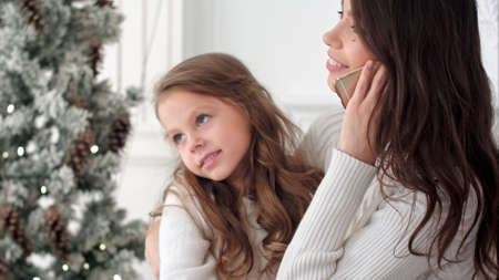Young mother talking on the phone with slightly bored daughter sitting on her lap next to the xmas treeの写真素材