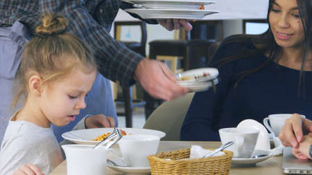 Cute little girl pouring cheese in her spaghetti in italian restaurant while her young parents have ate their vegetable salads yet.の写真素材